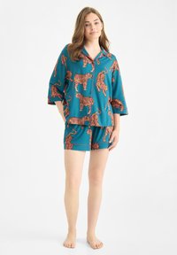 Mujer de pie descalza, vistiendo una camisa de pijama azul verdoso con botones y pantalones cortos con un estampado de tigre naranja sobre un fondo blanco.