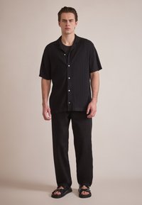 Svart kortermet skjorte med knapper og vertikal ribbing, kombinert med svarte løstsittende bukser og svarte sandaler med spenne detaljer.
