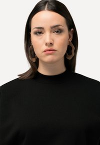 Suéter negro de punto con un cuello alto. El modelo lleva grandes pendientes ondulados de color marrón claro. Textura suave, diseño simple sin patrones.