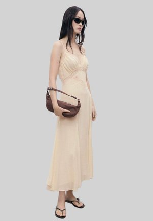 Light beige maxi dress with lace detailing, thin straps, and a flowing silhouette. Model carries a dark brown handbag and wears black sunglasses.