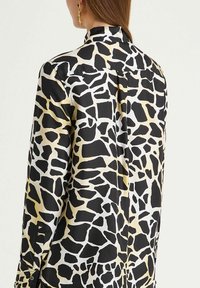 Woman wearing a black and beige giraffe-print long-sleeve blouse with a collar, shown from the back, with gold hoop earrings visible.