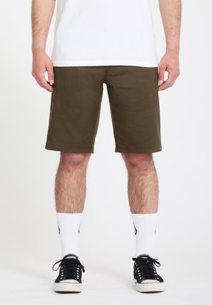 Homme portant un short vert olive, des chaussettes blanches montantes, des baskets noires et blanches, debout devant un fond blanc uni.