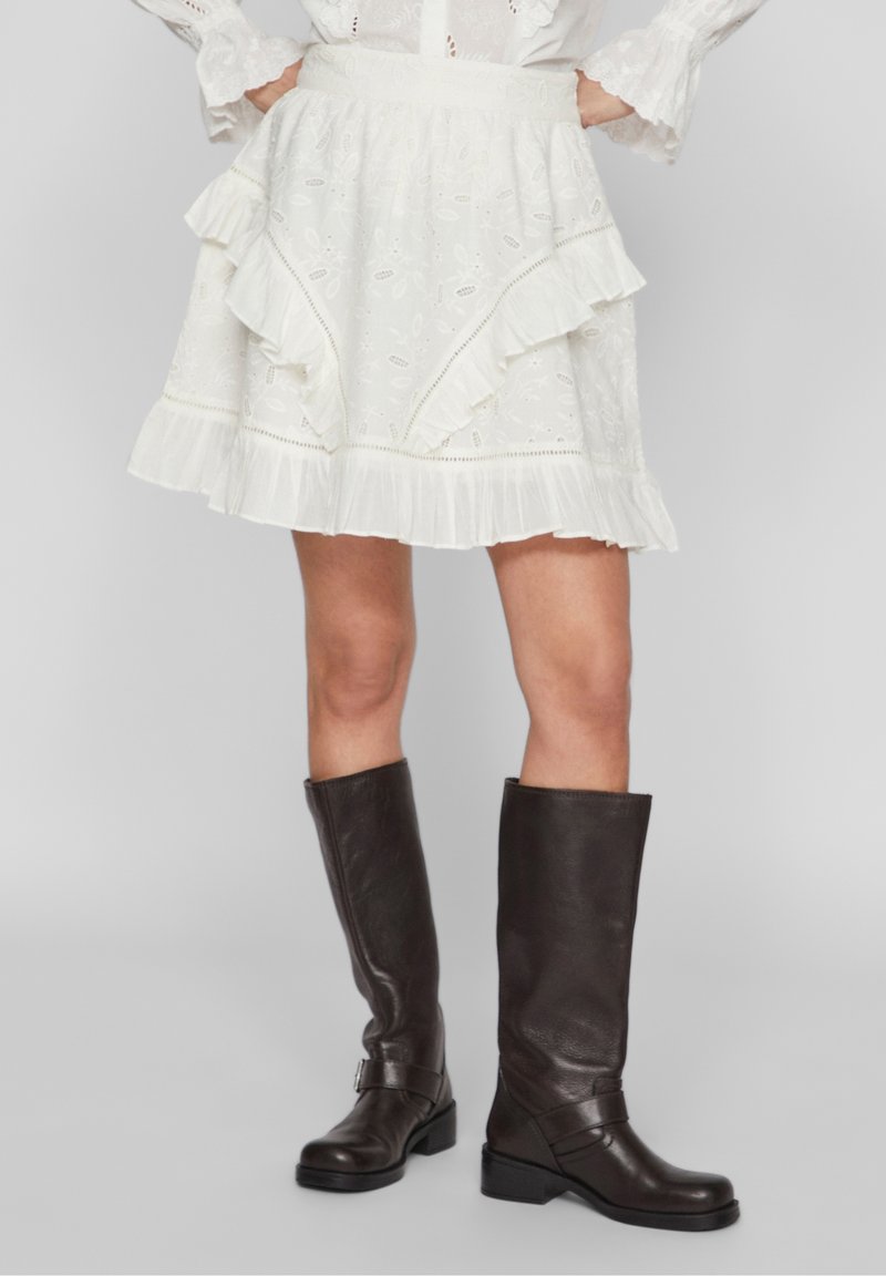 Žena oblečená v bílé sukni s volány a květinovou výšivkou, s koženými vysokými hnědými botami po kolena, stojící s rukama na bocích.
