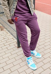 Pantaloni jogger viola con un logo di un toro rosso sulla coscia, indossati con sneaker azzurre e bianche, abbinati a una giacca di denim.