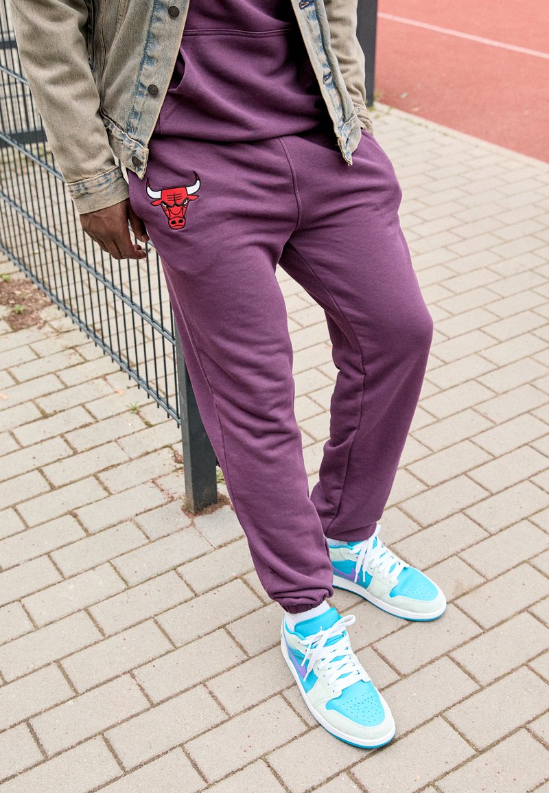 Pantaloni jogger viola con un logo di un toro rosso sulla coscia, indossati con sneaker azzurre e bianche, abbinati a una giacca di denim.
