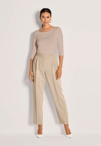 Vrouw met zwart haar in een strakke paardenstaart, gekleed in een beige top met driekwartmouwen, hoge beige broek, witte hakken en gouden ringoorbellen.