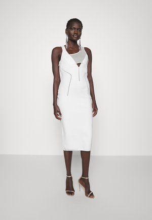 Fehér, testhez simuló ruha geometrikus mintával és kivágott részlettel a dekoltázson. Ezüst kiegészítők és magas sarkú cipők teszik teljessé a megjelenést.