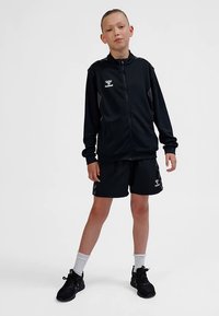 Zwart sportief jack met rits en logo, gecombineerd met zwarte korte broek. De outfit heeft een sportief ontwerp en wordt aangevuld met witte sokken en zwarte sneakers.