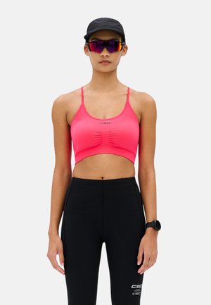 Frau trägt einen pinkfarbenen Sport-BH, schwarze hoch taillierte Leggings, schwarze Mütze, Sportsonnenbrille und eine schwarze Smartwatch, steht vor einem schlichten Hintergrund.