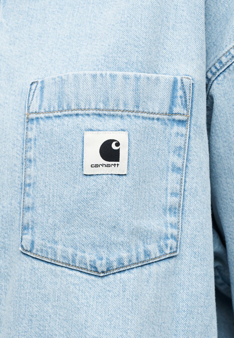 Light blue denim shirt featuring a square back pocket with a white label displaying the Carhartt logo in black. Visible stitching detail.