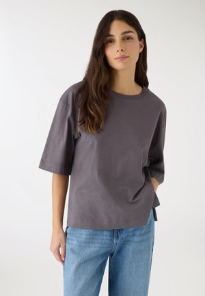 Giovane donna dai capelli castani lunghi indossa una maglietta grigio scuro ampia e jeans azzurri chiari, in piedi davanti a uno sfondo bianco uniforme.