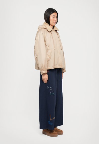Veste matelassée beige avec un col duveteux, associée à un pantalon large bleu marine arborant une broderie de texte colorée. Des chaussures à plateforme brunes complètent le look.