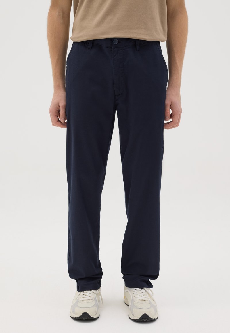 Pantalons en coton bleu marine avec une silhouette à jambe droite, fermeture par bouton à l'avant et poches latérales. Portés avec des baskets de couleur claire.