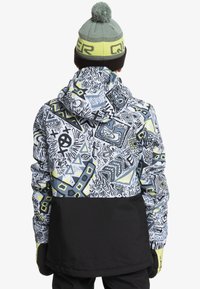 Veste de snowboard colorée avec des motifs géométriques en blanc, noir et accents jaunes, assortie à un bonnet vert avec un pompon sur le dessus.