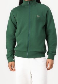 Sudadera verde con cremallera, hecha de tela suave, con cuello alto y logo de Lacoste en el pecho izquierdo; ajuste holgado con puños y dobladillo acanalados.