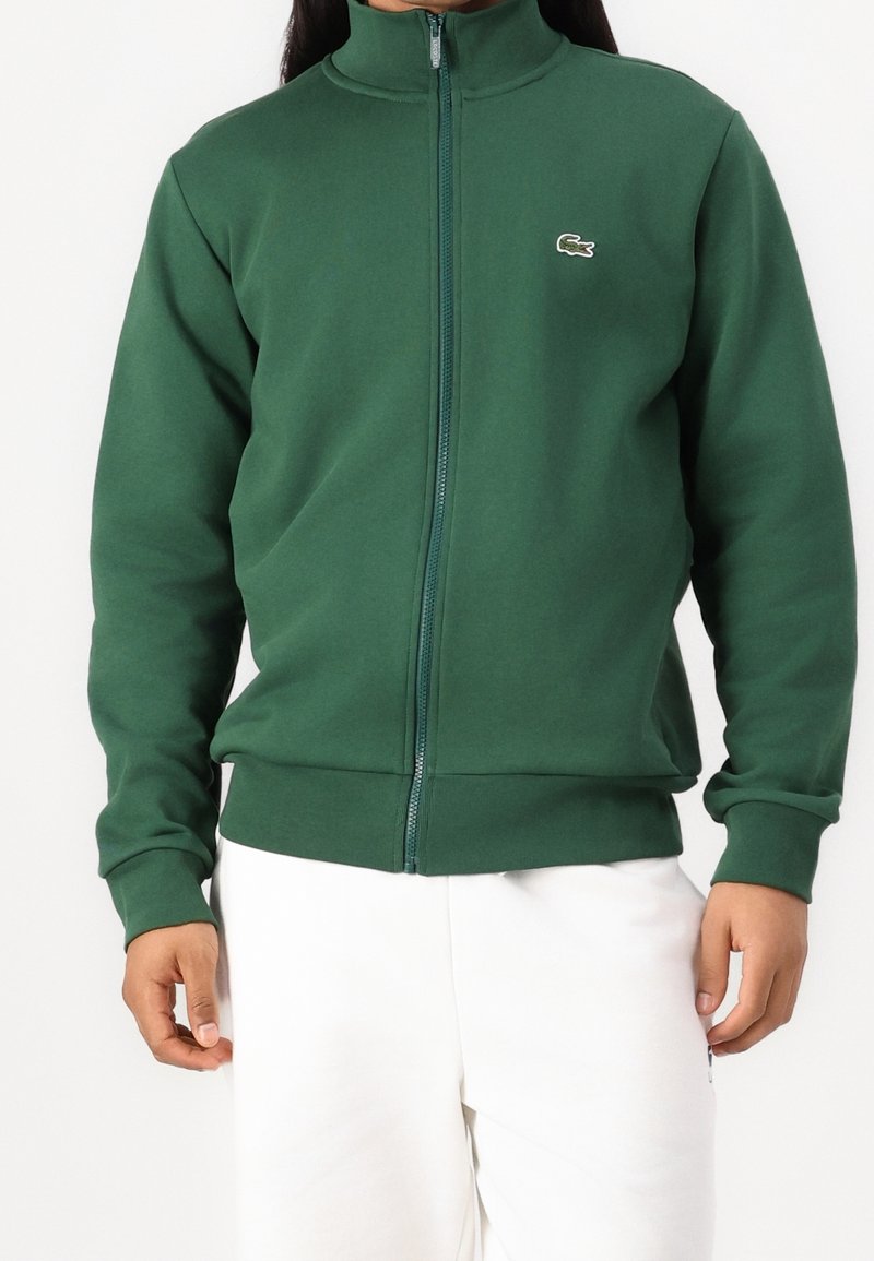 Sudadera verde con cremallera, hecha de tela suave, con cuello alto y logo de Lacoste en el pecho izquierdo; ajuste holgado con puños y dobladillo acanalados.