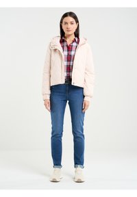 Gaiši rozā quiltēta jaka ar kapuci, elastīgām aprocēm un pogu aizdari, valkāta virs sarkana un zila ruļļa krekla un ziliem džinsiem.