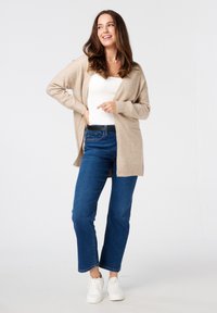 Beige gebreide cardigan met open voorkant, gedragen over een witte top. Paar met blauwe straight-leg jeans en witte sneakers. Zachte textuur is duidelijk zichtbaar.