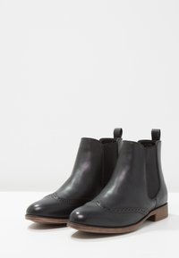 Stivaletti Chelsea in pelle nera con dettagli brogue, pannelli elastici ai lati e un tacco basso in legno, che mostrano una texture liscia e una forma classica.