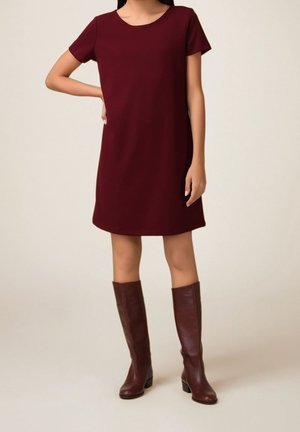 Kobieta ubrana w bordową sukienkę z krótkim rękawem, do której dobrała brązowe skórzane kozaki do kolan, stojąca na tle jednolitego tła.