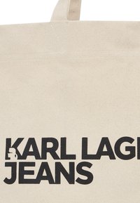 Bežinė drobinė rankinė su juodu blokuotu tekstu, kuris skaito "KARL LAGERFELD JEANS." Paprastas dizainas su tvirtais rankenomis.
