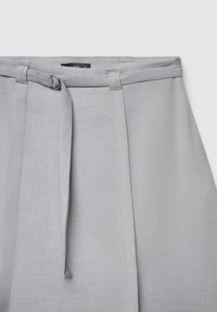 Pantalon gris clair avec une texture lisse, doté d'une taille ceinturée, de plis et d'un design épuré. Conçu pour une coupe structurée.