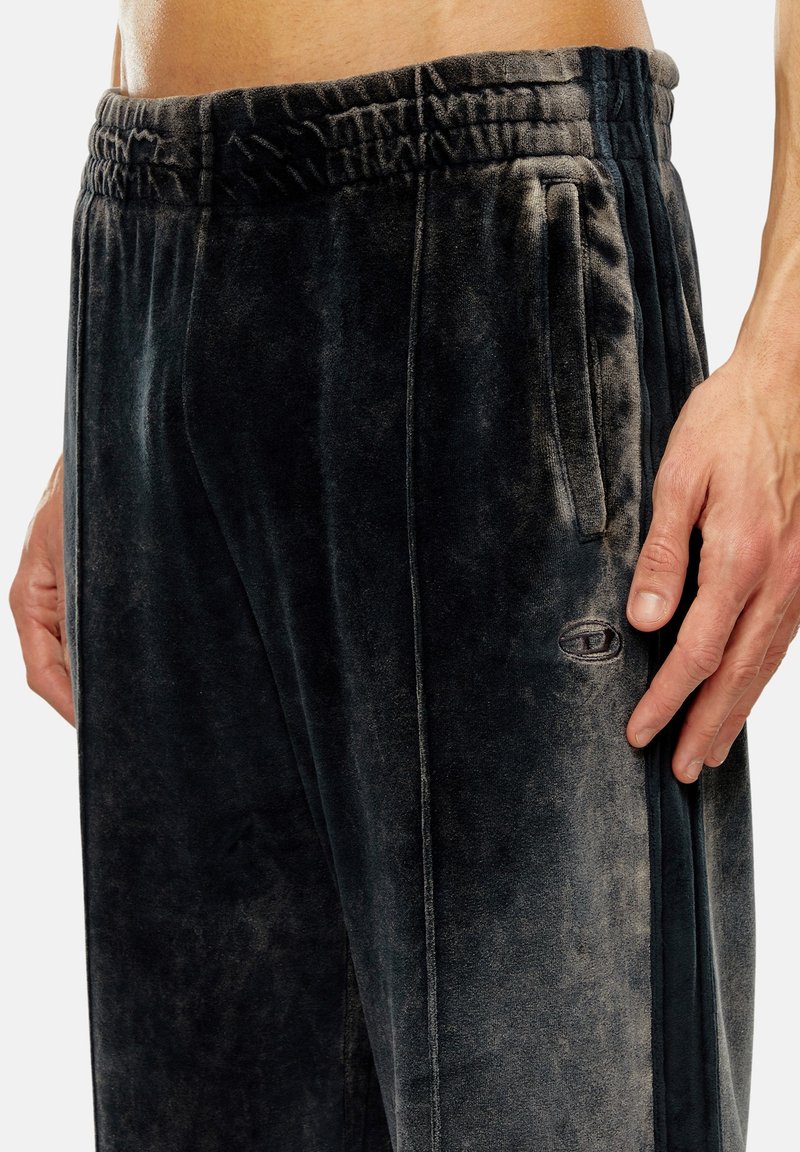 Nahaufnahme einer Person, die dunkelgraue Velours-Sweatpants mit elastischem Bund, seitlichen Taschen und einem kleinen ovalen Logo am rechten Oberschenkel trägt.