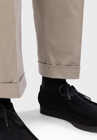 Beige Baumwollhosen mit umgeschlagenem Saum, kombiniert mit schwarzen Wildlederschuhen mit Schnürsenkeln und einer strukturierten Sohle. Sichtbare schwarze Socken.