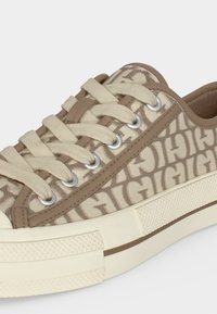 Baskets basses beige et taupe avec une tige en tissu présentant un motif de texte "GIGI" répété et des lacets beiges passés dans des œillets métalliques.