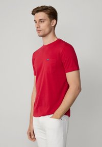 Rode katoenen T-shirt met korte mouwen, ronde halslijn en een borstzak aan de linkerkant. Voorzien van een klein blauw merkembleem op de zak.