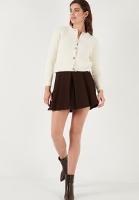 Cremefarbener, flauschiger Cardigan mit fünf glänzenden Knöpfen, kombiniert mit einem braunen Faltenrock und braunen Stiefeletten. Einfaches, elegantes Design mit weichen Texturen.
