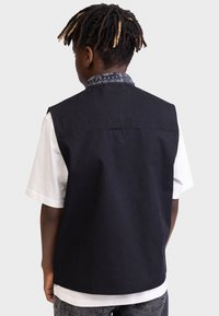 Gilet in denim nero senza maniche con colletto a motivo bandana, indossato sopra una maglietta bianca. Il retro presenta dettagli cuciti e un orlo curvo.