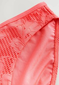 Korálové bikiny s dnom s kvetinovým čipkovaným vzorom na jednej strane a hladkým materiálom na druhej. Šité okraje a elastický pás.