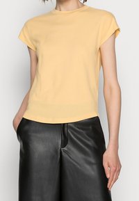 Donna che indossa una t-shirt a maniche corte di colore giallo chiaro, leggermente infilata nei pantaloni larghi in pelle nera a vita alta, con le mani nelle tasche.