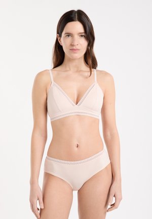 Femme aux longs cheveux bruns portant un soutien-gorge beige clair et une lingerie assortie, debout devant un fond blanc uni.