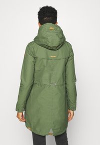 Olivgrön huva parkasjacka med dragsko i midjan, tryckknappar på ärmarna och ett horisontellt sömdekoration. Har en liten logotag.