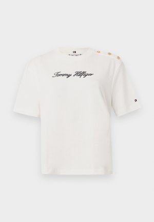 T-shirt en coton blanc à manches courtes, avec un logo noir "Tommy Hilfiger" en script et trois accents de boutons dorés sur l'�épaule gauche.