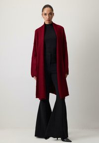 Nő hátranyalt hajjal, hosszú vörös kardigánt visel fekete garbó fölött, fekete bővülő nadrággal és hegyes orrú fekete magassarkúval.