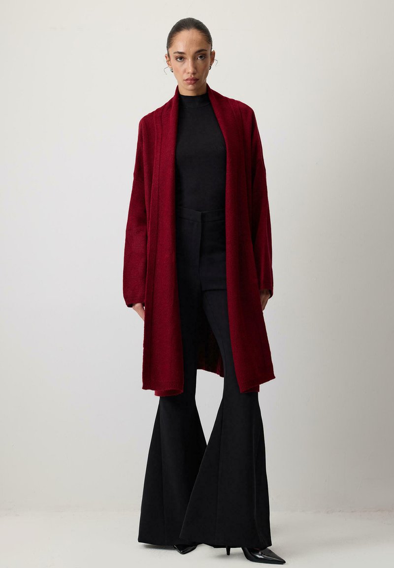 Nő hátranyalt hajjal, hosszú vörös kardigánt visel fekete garbó fölött, fekete bővülő nadrággal és hegyes orrú fekete magassarkúval.