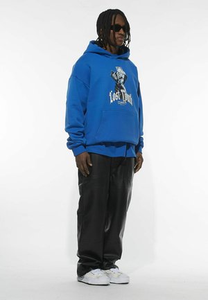 Blå hættetrøje med grafisk print, frontlomme, båret over en sort skjorte og sorte bukser i kunstlæder. Hvide sneakers fuldender outfittet.