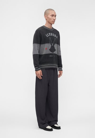 Homme portant un pull rayé noir et gris avec un motif de lapin et un pantalon large noir, debout devant un fond gris uni.