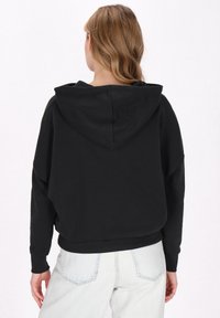 Felpa nera con cappuccio e vestibilità rilassata, con polsini e fascia in costina, vista da dietro, abbinata a jeans azzurri chiari.
