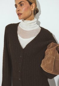 Brown knitted cardigan with a V-neck and ribbed texture, featuring button details. Worn over a white turtleneck and holding a tan suede jacket.