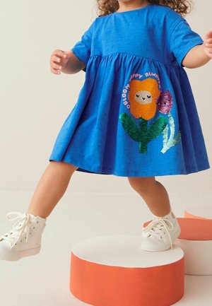 Småbarn i blå kjole med smilende blomstbroderi træder på orange og hvide cylindriske platforme iført hvide sneakers.