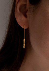 Goldene, längliche Ohrringe mit einer strukturierten rechteckigen Akzentuierung, die an einem dünnen, geraden Steg hängen. Die Oberfläche weist komplizierte Verzierungen auf.