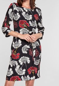 Robe florale avec une base noire, ornée de coquelicots blancs et rouges. Manches trois-quarts et coupe au genou. Tissu lisse avec un léger éclat.