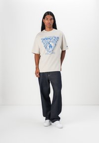 Beige kortærmet t-shirt med blå grafisk design, der viser teksten "Los Angeles Dodgers" og "siden 1958", parret med mørke denimbukser.