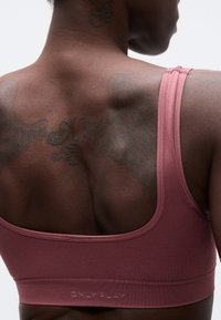 Κοντινό πλάνο πάνω μέρους της πλάτης ενός ατόμου με τατουάζ, που φοράει αθλητικό σουτιέν σε σκουρό ροζ χρώμα με το λογότυπο "ONLY PLAY" στη ζώνη.