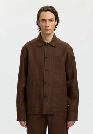 Młody mężczyzna o brązowych włosach do ramion, ubrany w zapinaną na guziki ciemnobrązową kurtkę z trzema przednimi kieszeniami na jednolitym szarym tle.