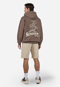 Brun hoodie med grafisk tryk af blade og ordet "SOMMER" på ryggen, parret med beige slidte shorts og sneakers.
